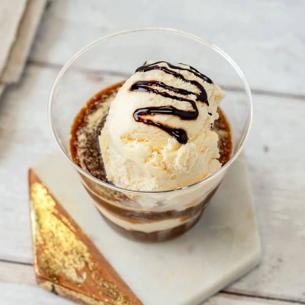 A clear glass of layered dessert with vanilla ice cream and chocolate drizzle on a marble coaster.