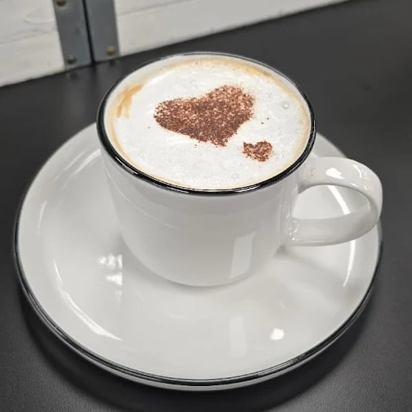 A white cup of coffee with a heart-shaped cocoa design on top of the foam, on a matching saucer.