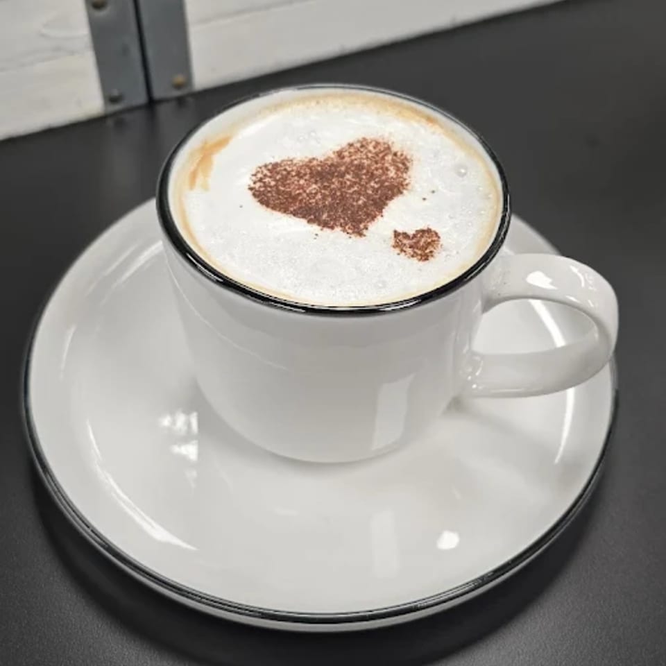 A white cup of coffee with a heart-shaped cocoa design on top of the foam, on a matching saucer.