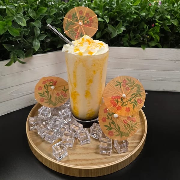 A mango smoothie with whipped cream and fruit pieces, garnished with paper umbrellas and ice cubes.