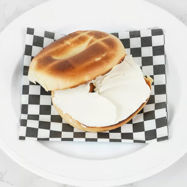Toasted bagel with a generous layer of cream cheese, served on a checkered paper on a white plate.